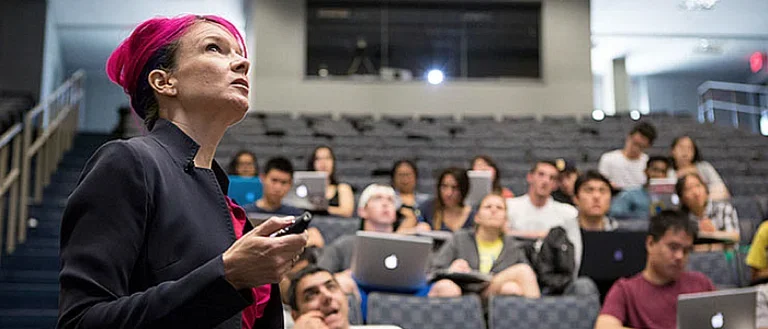 A researcher stands in front of an auditorium. Engaged students are seen to her right.