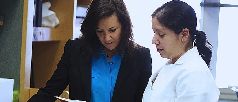 Two researchers, one in a black blazer and the other in a white labcoat, examine a page in a book.