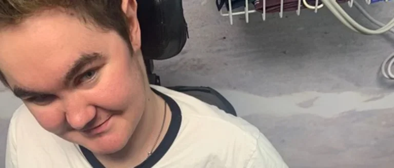 Young man with Duchenne Muscular Dystrophy wearing white shirt, smiling at camera. 