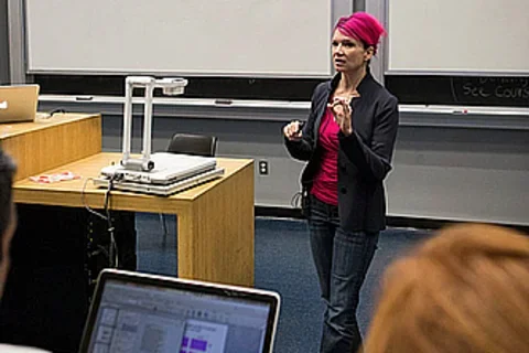 A lecturer stands at the front of an auditorium. A student's laptop is visible in the forefront.