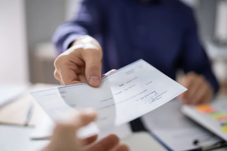 A business person's outstretched arm is seen handing a check to another person's outstretched arm.