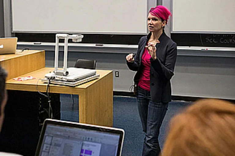 A lecturer stands at the front of an auditorium. A student's laptop is visible in the forefront.