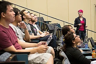 A lecturer stands at the far end of a lecture hall. Engaged students look ahead at a project screen.