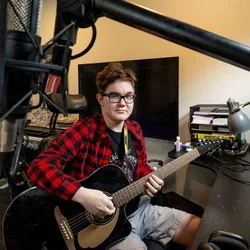 Cooper Jones in a red flannel shirt playing guitar in a studio.