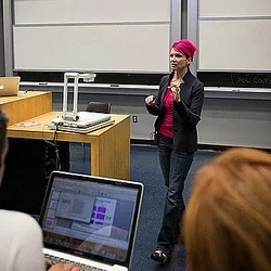 A lecturer stands at the front of an auditorium. A student's laptop is visible in the forefront.